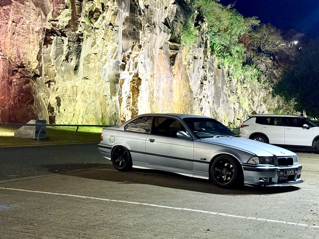 Silver BMW E36 coupe parked beneath an illuminated rock face at night